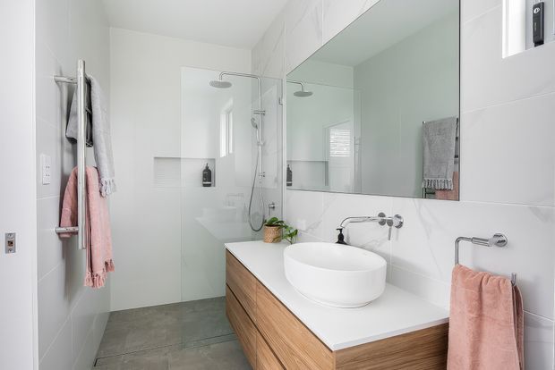Bright contemporary bathroom with a walk-in glass shower, wall-to-wall mirror, freestanding vessel basin, wall-mounted chrome tapware, and a floating timber vanity with a white benchtop. Large white wall tiles and grey stone-look floor tiles create a clean, light scheme, with soft pink towels adding warmth.
