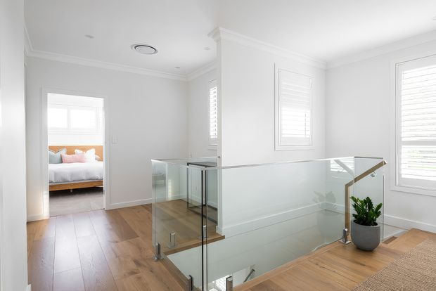 Bright contemporary upstairs landing with warm timber flooring, white walls and trim, plantation-shuttered windows, and a frameless glass balustrade around the stair opening. A bedroom is visible through the doorway, and a potted plant sits beside the railing.