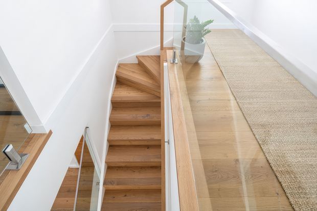 Bright contemporary stairway with warm timber treads and landing, white walls, and a clear glass balustrade framed in timber. A neutral woven runner covers part of the upper landing, with a potted plant adding a small natural accent. A mirror or reflective glazing at the lower left reveals additional stair and railing details.