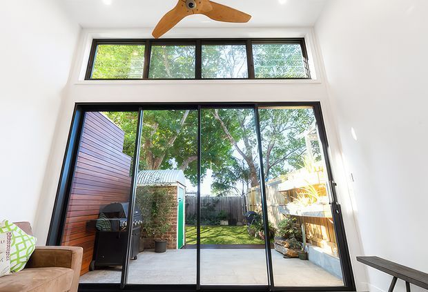 Bright interior view looking through large black-framed sliding glass doors to a landscaped courtyard and patio. The room has white walls, a high ceiling, clerestory windows, a timber ceiling fan, and partial living furniture. The glazing and courtyard connection dominate the image.
