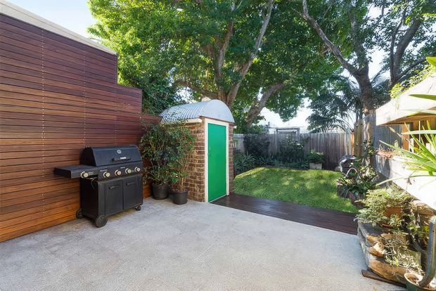 A sunny landscaped backyard patio with a large concrete-paved entertaining area, timber privacy screen, built-in barbecue, and a small brick outbuilding with a bright green door and curved corrugated roof. Timber decking leads through the garden, with mature trees, planting, and a timber boundary fence creating a private outdoor setting.