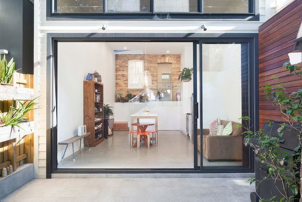 Bright contemporary view from an outdoor paved area through large black-framed glazed doors into an open-plan kitchen and living space. The interior features polished concrete flooring, white cabinetry, exposed brick detailing, timber shelving, and a compact dining table with warm timber accents. Exterior elements include dark horizontal timber cladding, rendered walls, and potted greenery.