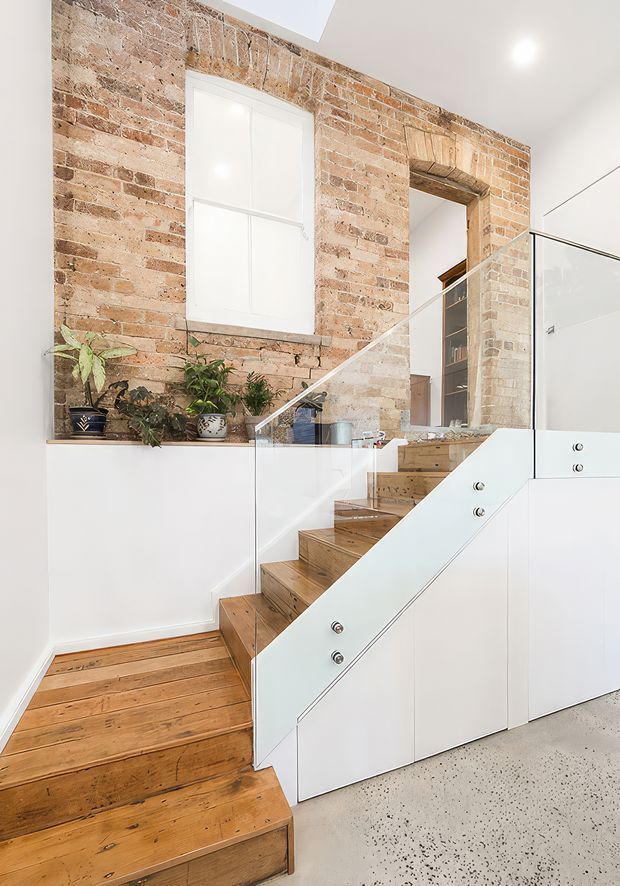 Bright stairway within a renovated interior, featuring an exposed warm brick wall, white built-in cabinetry beneath the stairs, timber stair treads and flooring, and clear glass balustrades. A small landing with potted plants and an upper-level doorway are visible. The space combines heritage industrial character with clean contemporary detailing.