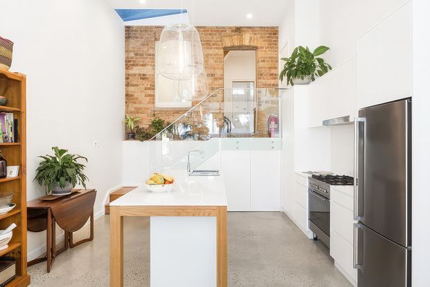 Bright open-plan kitchen with a white built-in kitchen along the right wall, stainless-steel refrigerator and gas range, a timber island with white benchtop, polished concrete flooring, and a glass balustrade leading to a raised mezzanine or stair area. Exposed warm brick walls, large pendant lights, indoor plants, and mid-century timber furniture add character. A skylight or roof glazing brings additional daylight into the high-ceilinged space.
