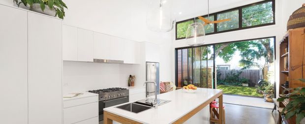 Bright contemporary kitchen with white flat-panel cabinetry, a large white island with integrated sink and timber edging, stainless steel range and appliances, and expansive black-framed glazing opening toward a leafy garden. Pendant lights, timber stools, indoor plants, and warm timber flooring add natural accents.
