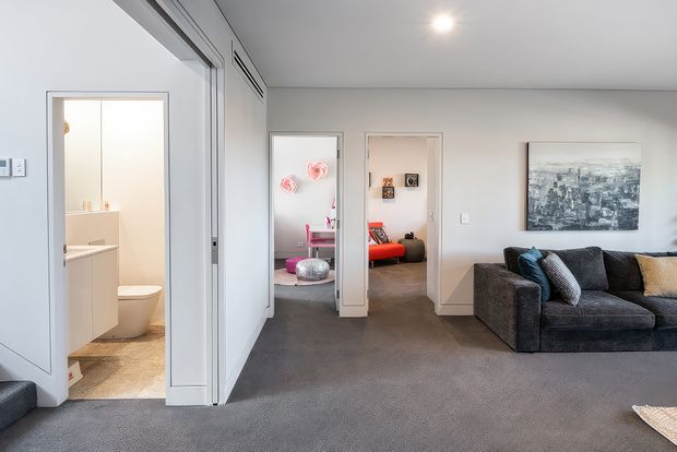 A contemporary living room with grey carpet, a large dark sectional sofa, framed wall art, and recessed ceiling lighting. Open doorways reveal a compact bathroom with a toilet and vanity, and a colourful adjoining room or bedroom with decorative furnishings. White walls, white door frames, and a broad glazed or mirrored sliding panel contribute to the bright, modern interior.