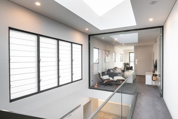 Bright contemporary interior landing or hallway overlooking a lower-level stairwell. The space features a large frameless glass balustrade, dark-framed horizontal louvre windows, a large skylight, recessed ceiling lights, pale walls, and grey carpet. A living area with lounge seating is visible beyond the glass partition, with a glimpse of the staircase below.