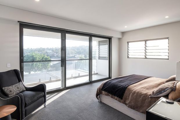 Bright contemporary bedroom with a large upholstered bed, dark bedding, a black leather armchair, carpeted floor, white walls and ceiling, and expansive black-framed sliding glass doors opening to a balcony. A horizontal louvered window provides additional ventilation and daylight.