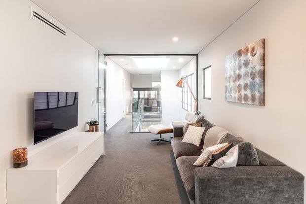 Bright contemporary living room with a large charcoal sectional sofa, white low-profile media console and wall-mounted television. The room opens toward a glazed stairwell and hallway, with recessed lighting, neutral walls, grey carpet, and a colourful circular wall artwork. A compact lounge chair and floor lamp sit near the staircase.