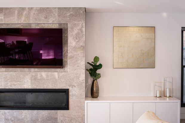 A refined contemporary living room feature wall with a large wall-mounted television above a linear fireplace, both set within a pale textured stone surround. A white built-in sideboard, sculptural artwork, decorative glass vessels, and a potted plant complete the calm neutral interior. Reflections in the television show part of the adjacent dining area.