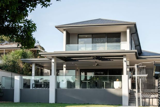 Contemporary two-storey residential house viewed from the rear, featuring a corrugated metal hipped roof, white rendered walls, extensive dark-framed glazing, a covered outdoor entertaining terrace with glass balustrade, and an upper-level balcony. The property has a clean monochromatic facade and landscaped garden surroundings.