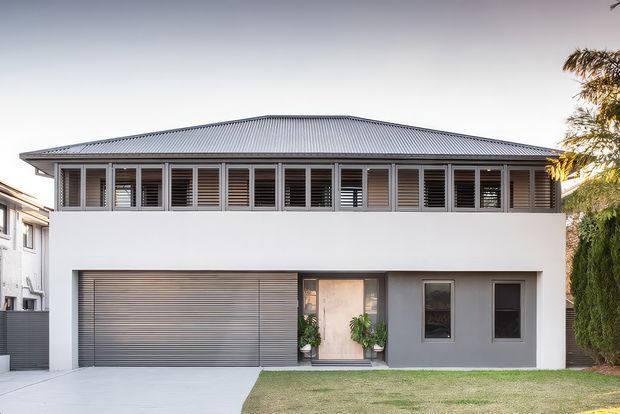 Front exterior view of a contemporary two-storey residence with a broad low-pitched corrugated metal roof, white rendered walls, a continuous upper-level band of timber-toned louvred windows, and a ground-floor garage and entry. The forecourt includes a concrete driveway and a small lawn, with neighbouring residential buildings and established planting at the edges.