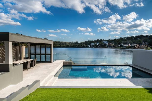 A contemporary waterfront outdoor entertaining area with a rectangular swimming pool in the foreground, pale tiled terraces, built-in outdoor kitchen or bar, and dining seating beneath a flat-roofed structure. Glass balustrades frame views across the calm water toward a residential shoreline. The scene is sunlit with a vivid blue sky and a clean, geometric architectural composition.