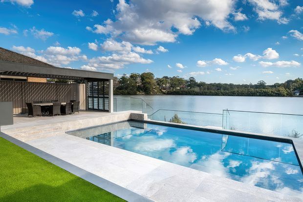 A contemporary waterfront outdoor entertaining area with a rectangular swimming pool overlooking a calm body of water. The pool is surrounded by pale stone paving, a frameless glass safety balustrade, and a covered terrace with outdoor dining furniture. The building has a low, flat-roofed form with dark vertical cladding and extensive glazing. The scene is bright and open under a blue sky.