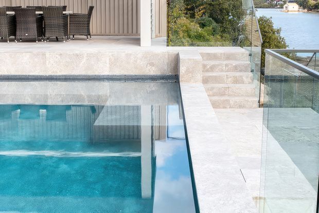 A contemporary outdoor pool area beside a waterfront, featuring a turquoise swimming pool, pale stone paving and steps, glass balustrades, and a covered outdoor dining area with woven furniture. The setting overlooks calm water and a landscaped shoreline.
