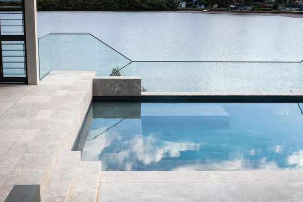 A contemporary waterfront pool terrace with a rectangular swimming pool edged in pale stone paving, a frameless glass balustrade, and a calm body of water extending beyond the terrace. Dark-framed glazing and a minimal architectural structure are visible at left, with a distant vegetated shoreline across the water.