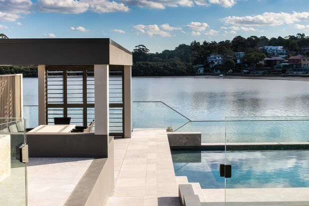 Contemporary waterfront terrace overlooking a calm body of water, with a geometric outdoor dining pavilion, pale stone paving, glass balustrades, and a swimming pool in the foreground. The scene features clean rectilinear forms, dark rendered walls, large glazed openings with horizontal screening, and a bright blue-sky setting.
