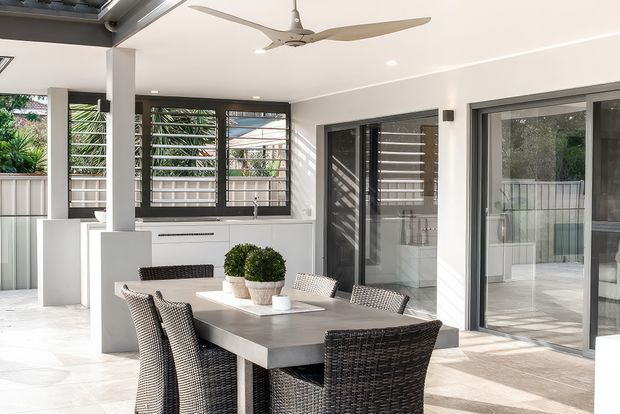 Bright contemporary dining and entertaining area with a large rectangular dining table and woven chairs, opening onto a covered outdoor terrace through expansive sliding glass doors. The space features white walls and ceiling, dark aluminium-framed glazing, louvred windows, pale tiled flooring, integrated white cabinetry, and a ceiling fan.