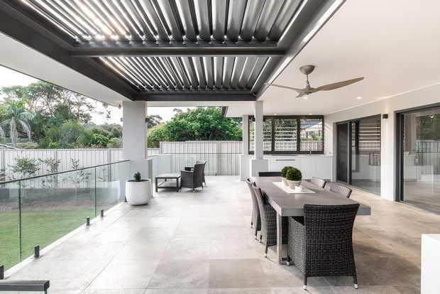 A spacious covered outdoor entertaining area with a dining table and wicker chairs, adjoining a glazed interior space. The terrace features large pale tiled flooring, glass balustrades, a louvred pergola roof, ceiling fan, built-in barbecue or cabinetry area, and views to a landscaped garden.