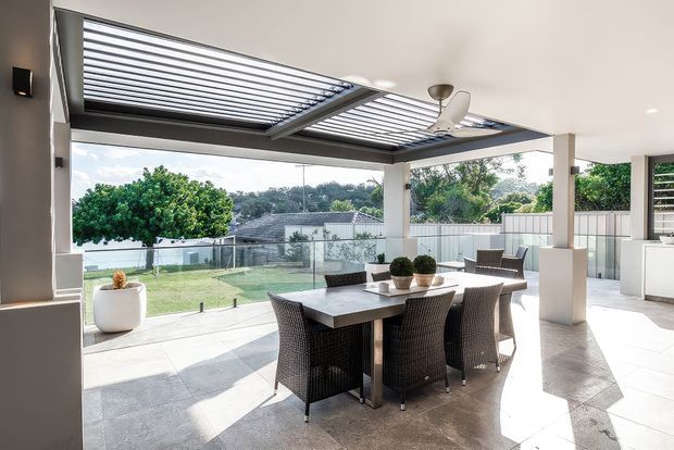Bright covered outdoor entertaining area with a dining table and woven chairs, opening onto a glass-balustraded terrace and suburban garden. The space features large retractable louvred roof panels, white structural columns, tiled flooring, and a ceiling fan, with an adjacent outdoor kitchen or service area visible at right.