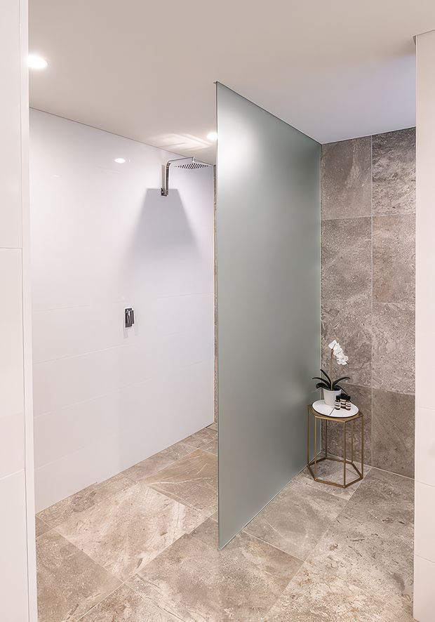 Contemporary walk-in shower area with a frameless frosted-glass privacy screen, large-format pale stone-look tiles on the floor and feature wall, white wall tiles, recessed lighting, and chrome shower hardware. A small metal side table with minimal decor sits beside the screen.