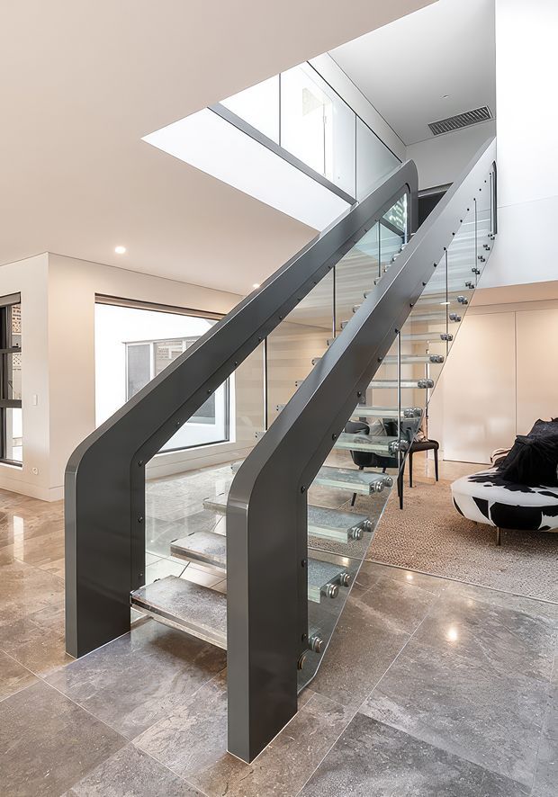 Bright contemporary interior centred on a sculptural open-riser staircase with dark metal side stringers, clear glass balustrades, and transparent tread supports. The staircase rises through a large skylight or void, with pale walls, recessed lighting, polished stone-look flooring, and a partially visible living area with a patterned rug and curved seating.