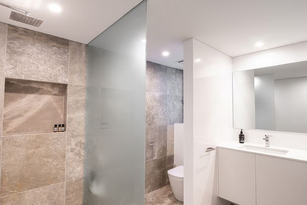 Contemporary bathroom with a walk-in shower enclosed by a frosted glass screen, large textured stone-look wall and floor tiles, recessed shower niche, wall-hung toilet, and a minimalist white floating vanity with integrated basin and large mirror. Recessed ceiling lighting creates a bright, clean atmosphere.
