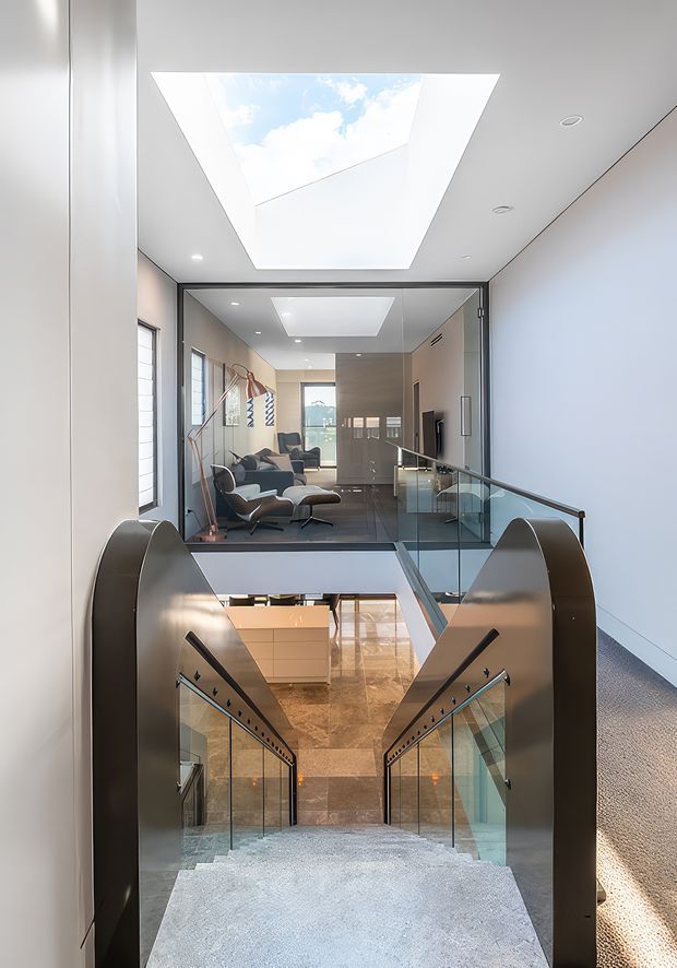 A bright, modern multi-level interior centered on a broad staircase descending through an open stairwell. The staircase has glass balustrades, dark metal sculptural side rails, and pale carpeted steps. A polished stone or marble landing is visible below, while a glazed living area with contemporary lounge furniture sits beyond the upper landing. Large skylights, recessed lighting, white walls, and extensive internal glazing create an airy, light-filled composition.