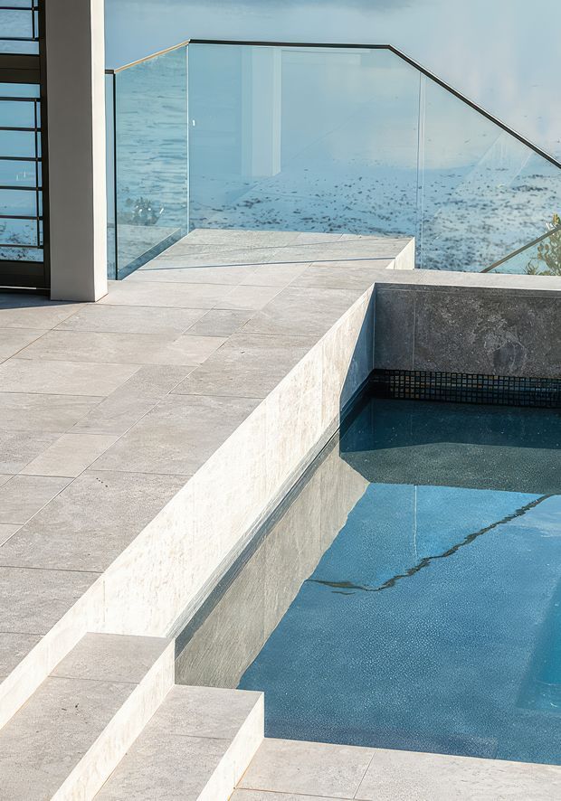 A contemporary outdoor terrace beside a swimming pool, finished with large pale stone-look paving and matching rendered pool walls. Frameless glass balustrades overlook a bright coastal or water view, while a dark tiled waterline and integrated pool steps add contrast. The image emphasizes clean geometric forms, layered levels, and the relationship between the terrace and pool.