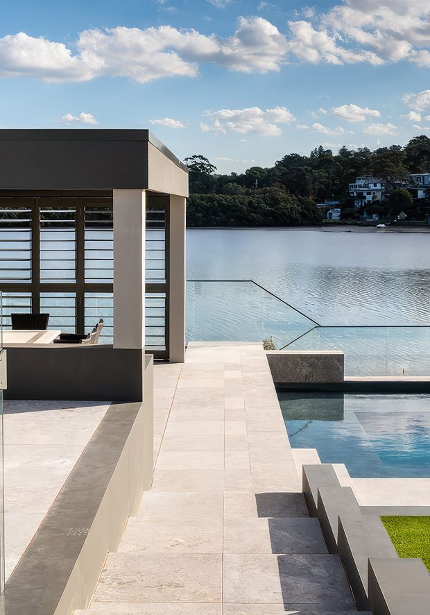 Modern waterfront residential terrace with pale stone paving, angular concrete or rendered walls, a reflective swimming pool, and frameless glass balustrades overlooking a calm lake. Floor-to-ceiling horizontal-louvered glazing encloses a dining area, while a neighbouring wooded shoreline and houses are visible across the water.