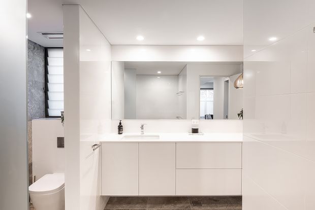 Bright, compact contemporary bathroom vanity area with a long white floating cabinet, integrated white benchtop and basin, large wall-to-wall mirror, glossy white wall finishes, recessed ceiling lights, and grey floor tiles. A toilet and shower area are visible to the left, while a doorway and reflected cabinetry appear in the mirror.