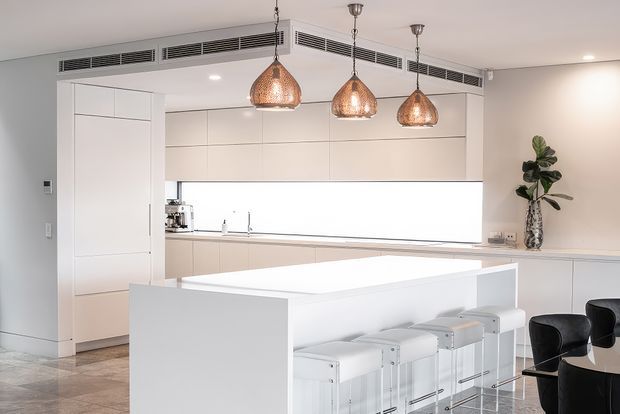 Bright, contemporary open-plan kitchen with extensive white flat-panel cabinetry, a large white island with integrated seating, a long illuminated splashback, and three woven copper pendant lights. Black dining chairs and a decorative plant add contrast, while stone-look flooring and concealed linear air-conditioning vents complete the clean, refined fit-out.