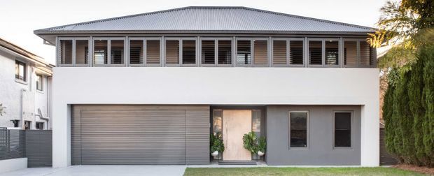 Front exterior view of a contemporary two-storey residence with a broad low-pitched metal roof, white rendered upper walls, grey lower-level facade, a wide louvred window band, roller garage door, and recessed entry. The home sits in a suburban setting with neighbouring structures and narrow landscaping visible at the edges.