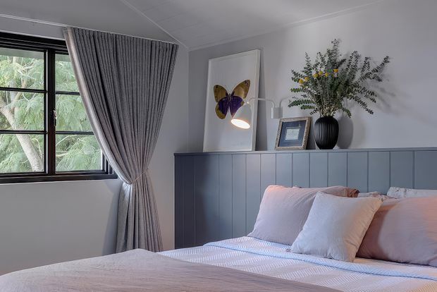 A softly styled bedroom with a pale upholstered bed, layered white and blush bedding, and a blue-grey vertical-panel headboard wall. A black-framed window with pleated grey curtains overlooks greenery, while a slim floor lamp, framed butterfly artwork, and a dark vase with foliage sit on the built-in headboard ledge.