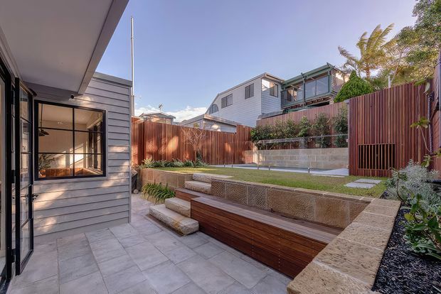 A landscaped residential courtyard viewed from a paved patio, featuring tiered garden beds, rendered retaining walls, timber screening and fencing, built-in timber bench seating, and a contemporary grey weatherboard extension with black-framed glazing. The sunny outdoor space has a warm, modern coastal-suburban character.