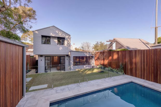 Exterior view of a compact two-storey home with grey horizontal weatherboard cladding, dark-framed windows, a small covered outdoor living area, timber privacy fencing, landscaped lawn and a rectangular swimming pool in the foreground. The property sits in a suburban residential setting with neighbouring rooflines visible.
