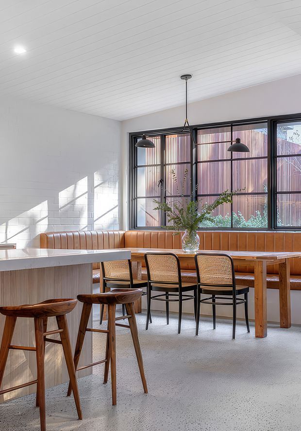 Bright contemporary dining and kitchen area with a built-in tan leather banquette, timber dining table, black-framed industrial-style windows, woven-back chairs, and pendant lighting. The space features white painted brick walls, a white-lined ceiling, and a polished light terrazzo or concrete floor, with abundant natural light.