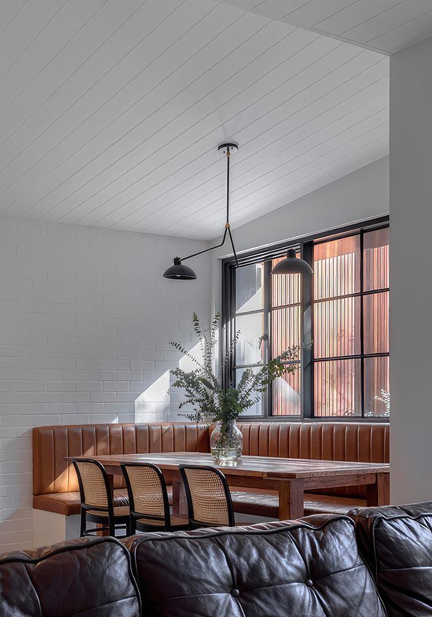 A stylish dining area with a built-in caramel-brown leather banquette wrapping around a timber dining table, paired with cane-backed black dining chairs. The space features white painted brick walls, a white linear-panel ceiling, black pendant lights, and large industrial-style black-framed windows with vertical screening. A dark leather sofa occupies the foreground, creating a warm restaurant or hospitality-inspired atmosphere.
