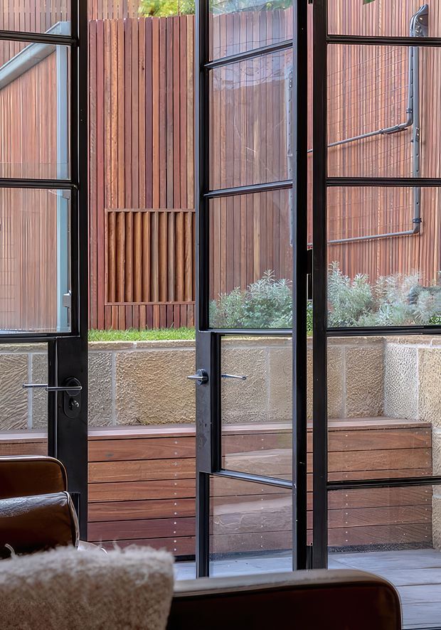 Interior view through tall black-framed glazed doors and windows toward a compact landscaped courtyard. The courtyard features vertical timber screening, a textured stone or concrete retaining wall, timber decking and built-in timber benching, with a glimpse of leather seating in the foreground. The composition emphasizes the contrast between dark metal frames, warm timber cladding and the enclosed outdoor space.