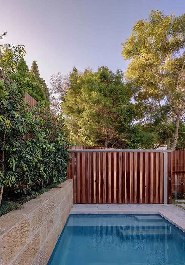 A landscaped residential pool courtyard with a narrow blue swimming pool in the foreground, surrounded by pale grey paving. A tall reddish-brown vertical timber slat fence and matching gate enclose the space, while dense mature greenery rises behind a textured concrete block retaining wall. The image has a warm, tranquil outdoor atmosphere.