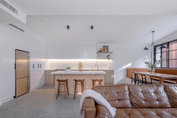 Bright open-plan kitchen and living area with a large central island, white flat-panel cabinetry, integrated stainless-steel refrigerator, timber accents, and three timber bar stools. A brown leather sofa occupies the foreground, while a dining table sits beside tall glazed doors with dark frames. The room has a white-lined ceiling, polished concrete or terrazzo-like flooring, and abundant natural light.