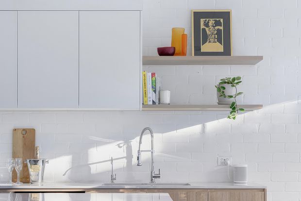 Bright, minimalist kitchen vignette with white painted brick walls, pale flat-panel overhead cabinetry, open timber shelves, a light stone benchtop, and a stainless-steel gooseneck tap. Decorative glassware, books, a framed artwork, and a trailing plant add subtle colour and texture.