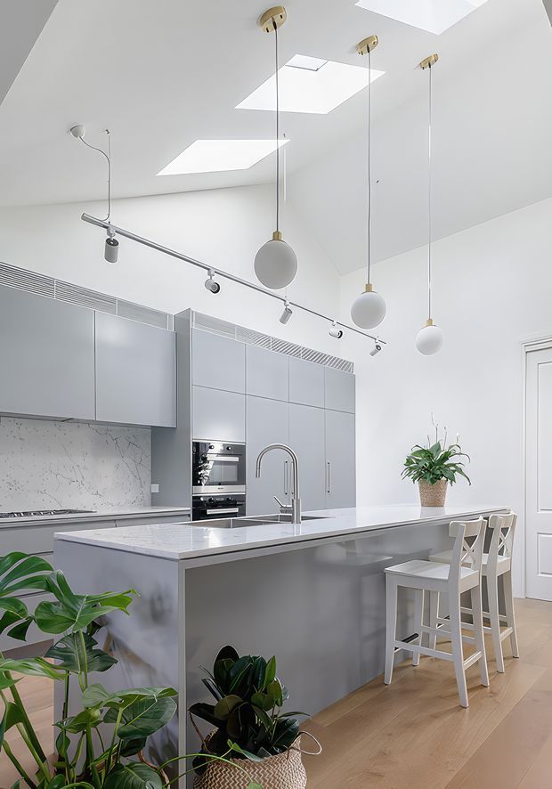Bright contemporary kitchen with a large grey island, integrated cabinetry and appliances, pale stone or composite benchtops, a light marble-look splashback, timber flooring, and white walls. Three spherical pendant lights hang above the island, with additional track lighting and multiple skylights. White bar stools and indoor plants add contrast.
