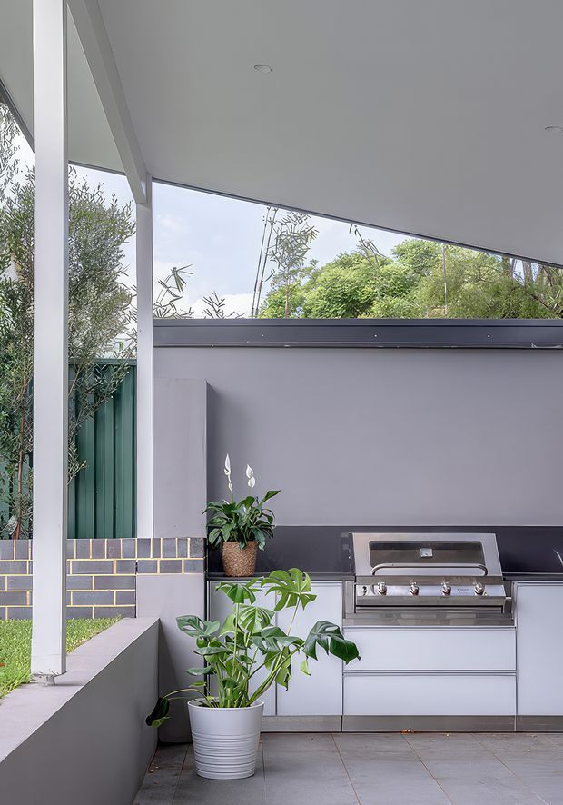 A covered outdoor kitchen and barbecue area with a stainless-steel built-in grill, pale cabinetry, dark tiled splashback, and a grey rendered wall. Large openings frame greenery, while potted tropical plants and a high angled ceiling create a bright, sheltered entertaining space.
