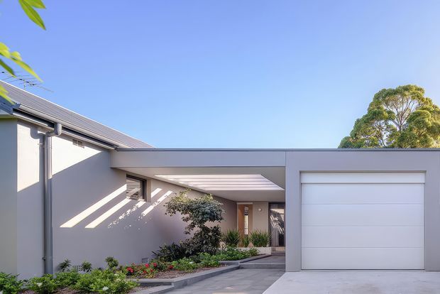 Contemporary single-storey residential facade with a pale rendered exterior, low-pitched metal roof, broad flat-roofed connecting canopy, and a large white sectional garage door. A paved driveway leads toward a recessed entry, bordered by established garden planting. The image is photographed outdoors in bright daylight with a clear blue sky.