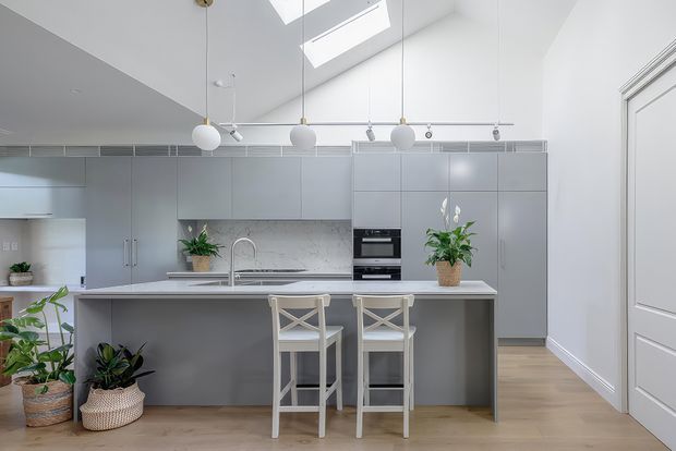 Bright contemporary kitchen with a large central island and two cross-back bar stools. The room features smooth pale grey full-height cabinetry, a light stone-look benchtop and splashback, integrated appliances, timber flooring, white walls, pendant globe lights, and a roof skylight. Plants add natural accents, while a detailed white internal door and high ceiling contribute architectural character.