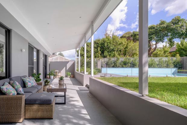 A covered outdoor living veranda runs alongside a modern home, with grey cushioned wicker seating, coffee tables, potted plants, concrete flooring, and slender white structural posts. The open side overlooks a landscaped lawn and rectangular swimming pool, with trees and a bright sky beyond.