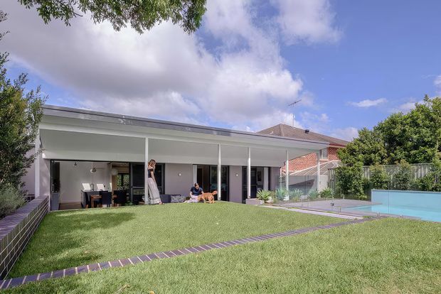 Exterior view of a contemporary single-storey home with a broad covered patio, slim white columns, extensive glazing, a large lawn, paved walkways, and a rectangular swimming pool. A pitched terracotta-tiled roof from the neighbouring or original house is visible behind the modern extension.