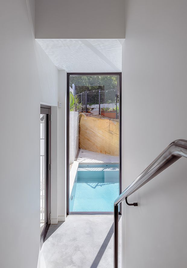 Bright minimalist stairway or landing with white walls, a pale concrete or stone floor, and a dark-framed full-height glass door overlooking a turquoise swimming pool and landscaped courtyard. A sleek dark metal handrail follows the stairs in the foreground, while strong sunlight creates crisp shadows and highlights the vertical architectural volume.