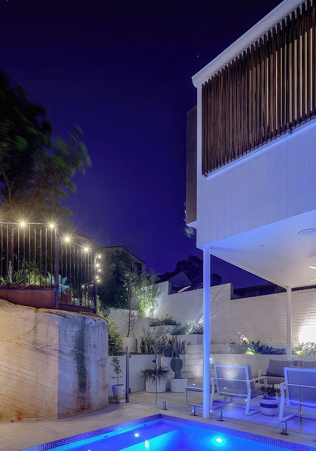 Night-time architectural photograph of a contemporary outdoor courtyard and pool area. The scene features a white multi-level building with a covered patio, vertical timber screening, white brick walls, outdoor dining/lounge furniture, landscaped planters, decorative lighting, and a brightly illuminated swimming pool in the foreground.